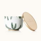 A gold-lidded candle tin with a white body and green Christmas tree patterns. The lid has a gold finish and the brand 'Thymes' is visible on it. Available at Be Charmed.  