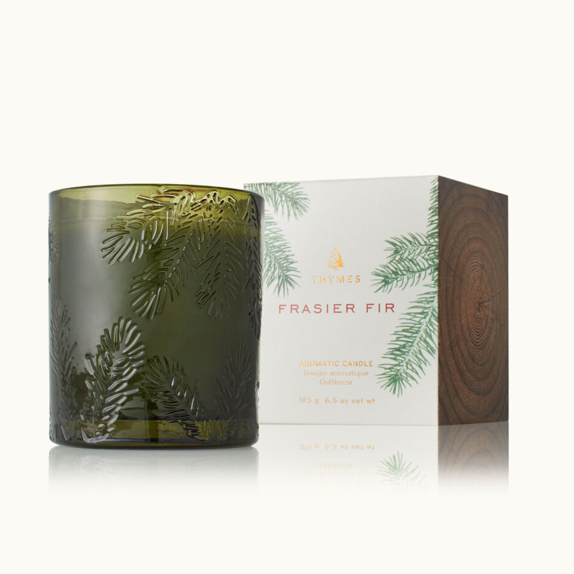 A green glass candle with a Frasier Fir pine needle design, accompanied by a white box with 'Thymes' and 'Frasier Fir' text on it. Available at Be Charmed