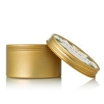 A gold travel tin candle with a lid, featuring a label with a Christmas tree and snowflake design. Available at Be Charmed