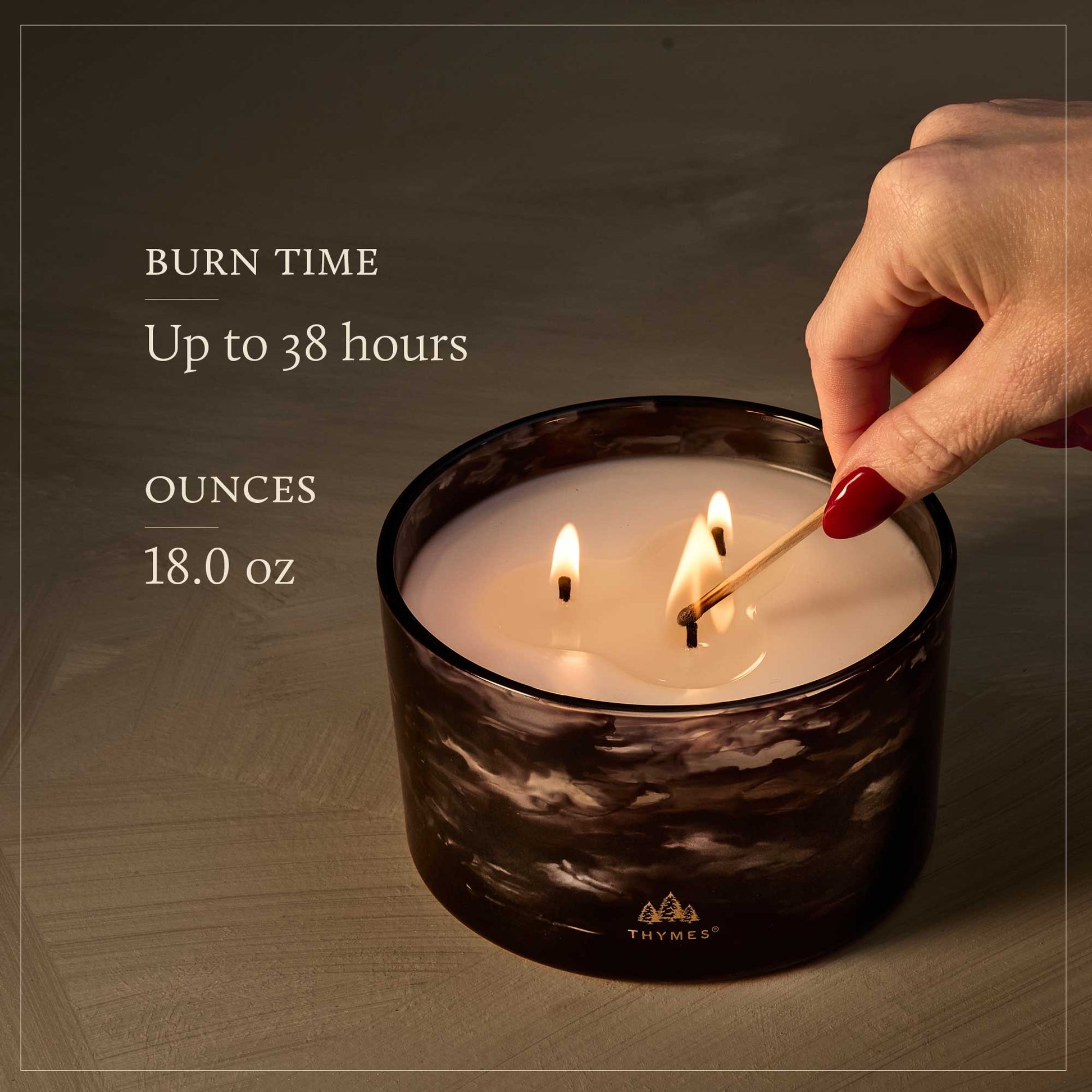 Thymes Noir Woods candle being lit with text about burn time and ounces, on a dark background. Available at Be Charmed.  
