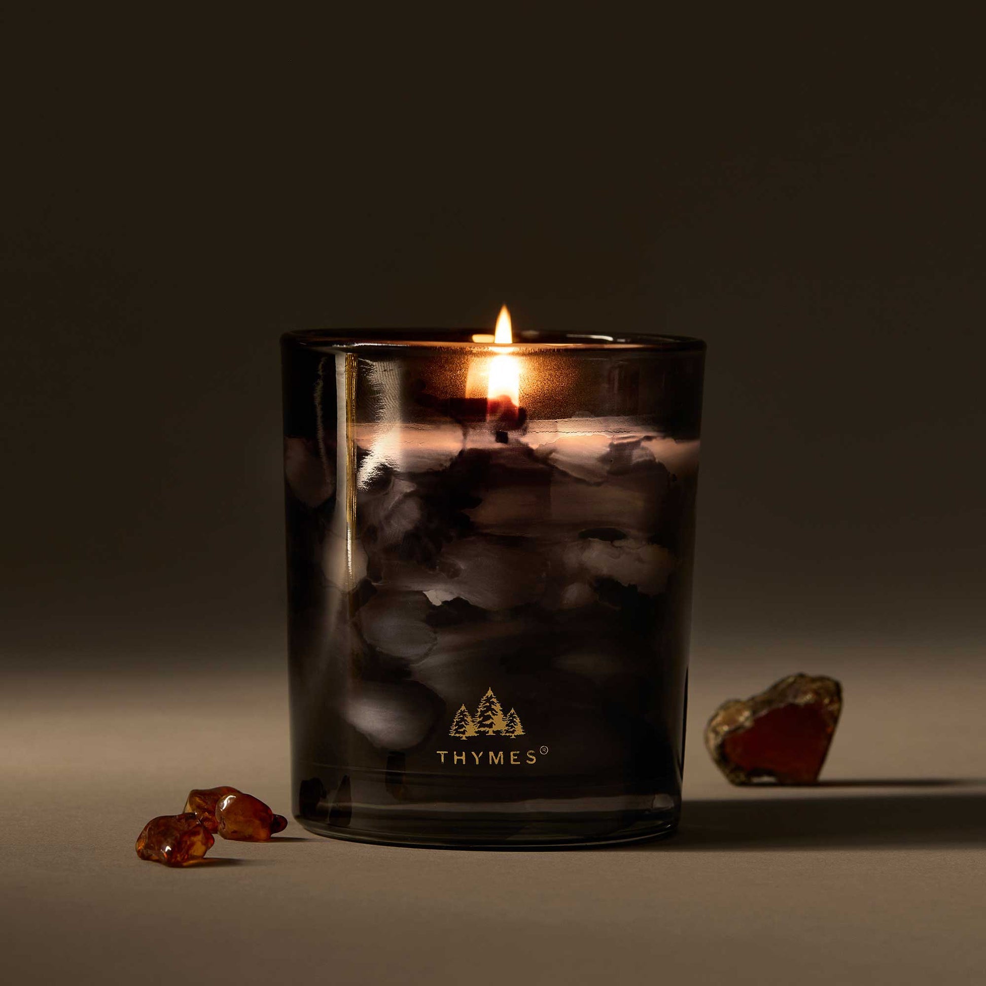 Thymes candle in a dark glass container with a lit candle inside, surrounded by amber-colored stones on a dark background. Available at Be Charmed.  