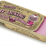 A gold-colored metal lip balm tin with a pinkish-bubble gum-scented lip balm inside. The lid features vintage-style graphics including a cup with a bubble and the text 'Vintage Lip Licking Bubble Gum'.Available at Be Charmed