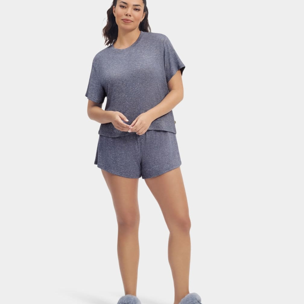 A woman standing and wearing a navy heather gray UGG Aniyah sleepwear set, consisting of a boxy fit tee and matching sleep shorts with a ruffle waistband. Available at Be Charmed