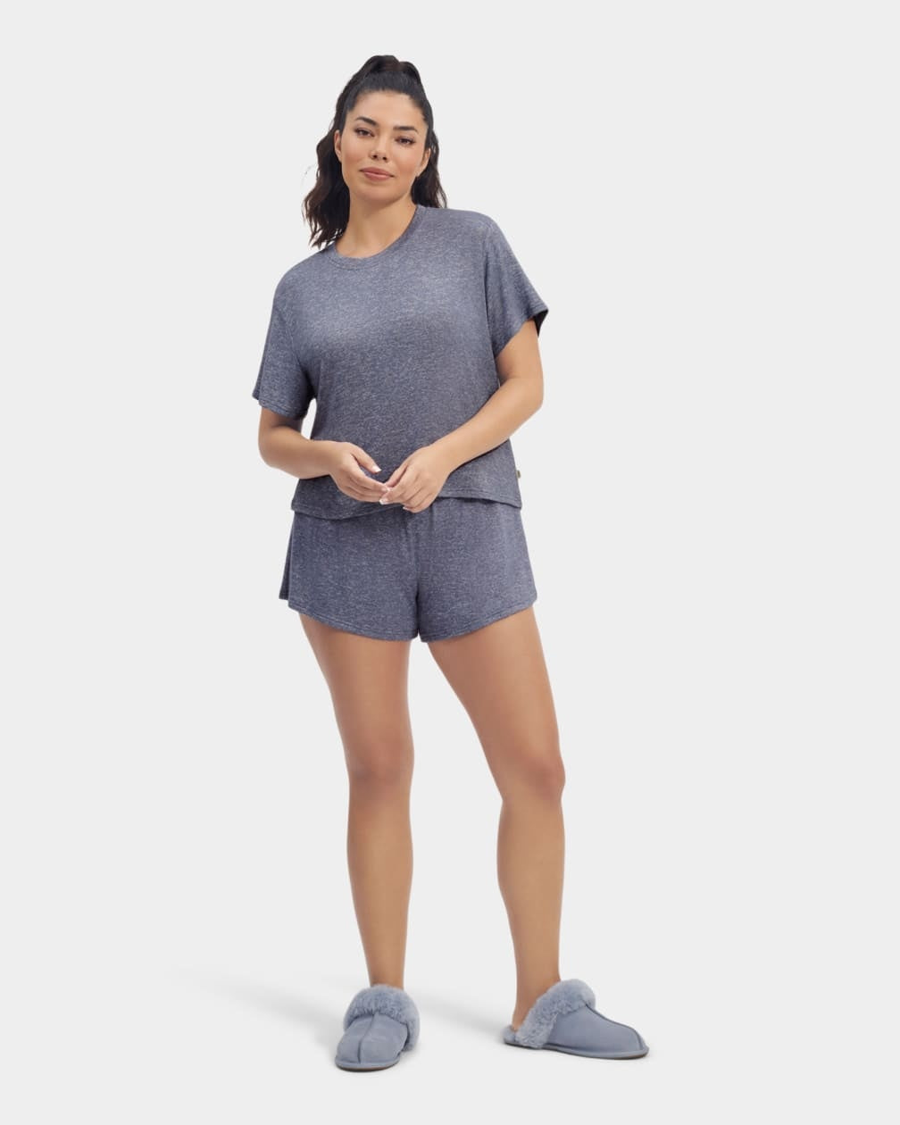 A woman standing and wearing a navy heather gray UGG Aniyah sleepwear set, consisting of a boxy fit tee and matching sleep shorts with a ruffle waistband. Available at Be Charmed