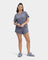 A woman standing and wearing a navy heather gray UGG Aniyah sleepwear set, consisting of a boxy fit tee and matching sleep shorts with a ruffle waistband. Available at Be Charmed