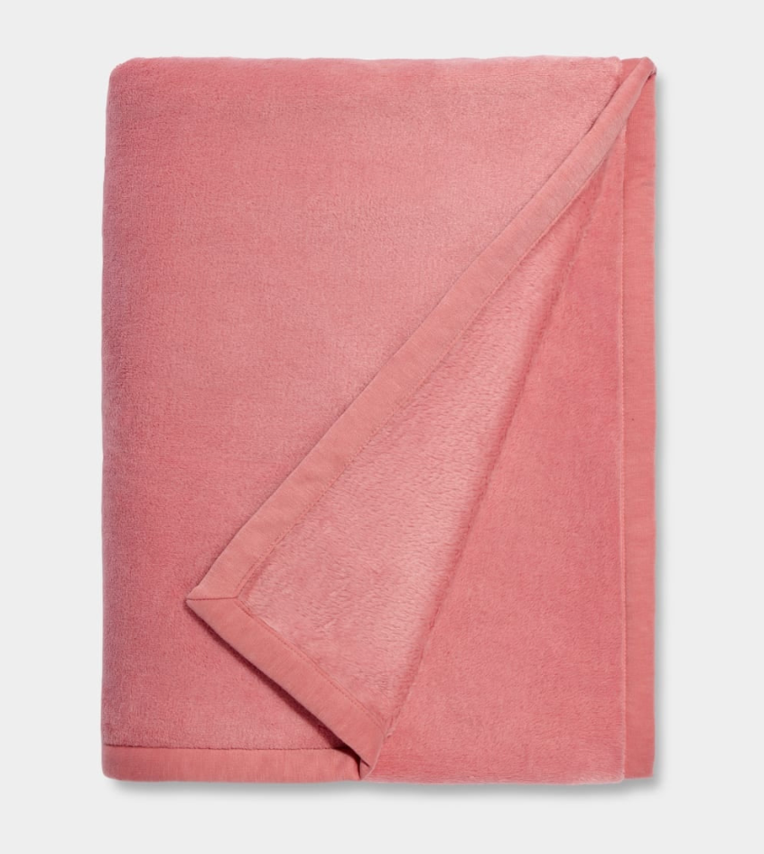 A pink UGG Duffield II throw blanket with a subtle leather-like finish on the edges, folded loosely. Available at Be Charmed