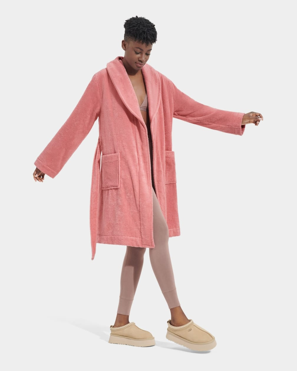 A model wearing a pink UGG Lenore Terry Robe with traditional patch pockets and an embroidered UGG heritage logo. Available at Be Charmed