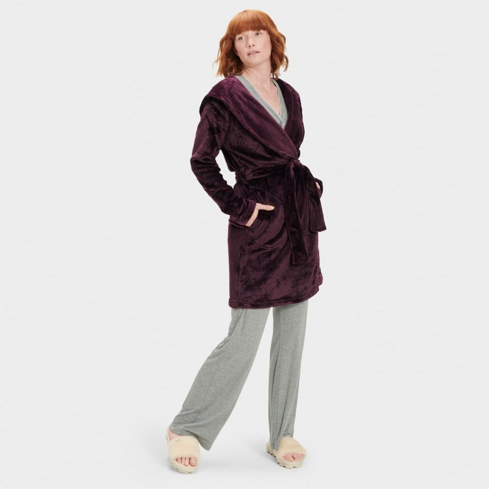 A woman standing and modeling a purple UGG robe with a neutral facial expression, wearing matching pants, with her hands in pockets. Available at Be Charmed