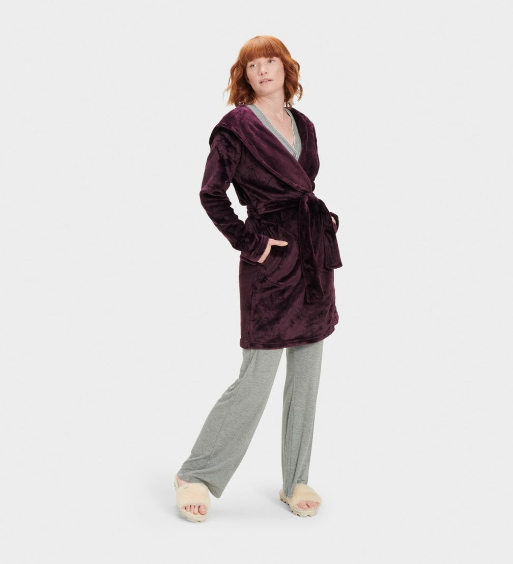 A woman standing and modeling a purple UGG robe with a neutral facial expression, wearing matching pants, with her hands in pockets. Available at Be Charmed