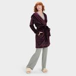 A woman standing and modeling a purple UGG robe with a neutral facial expression, wearing matching pants, with her hands in pockets. Available at Be Charmed