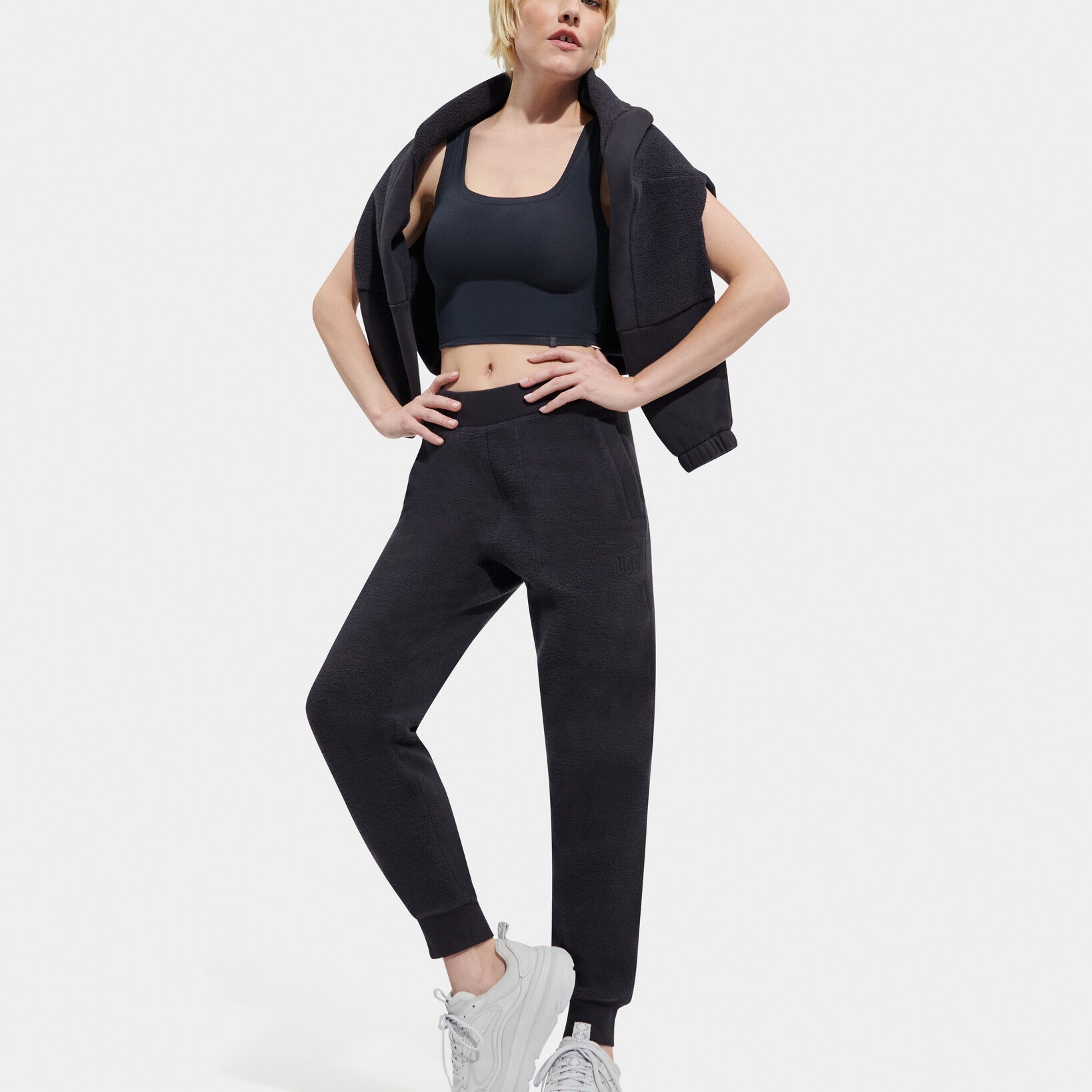 A female model standing, posing, and turning to face the camera. She is wearing a black UGG Ink Sofiana Mixed Jogger with an elastic waistband, and white sneakers. Available at Be Charmed