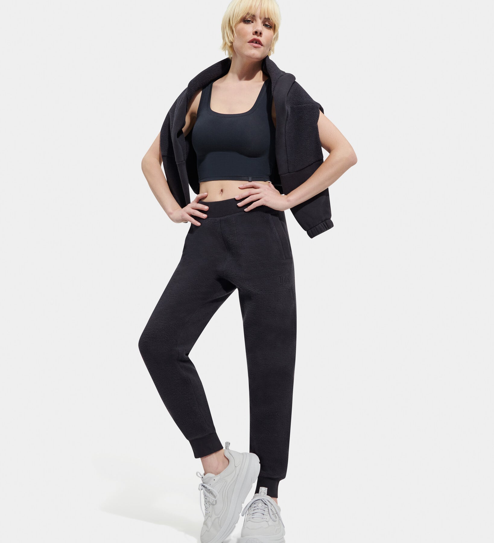 A female model standing, posing, and turning to face the camera. She is wearing a black UGG Ink Sofiana Mixed Jogger with an elastic waistband, and white sneakers. Available at Be Charmed