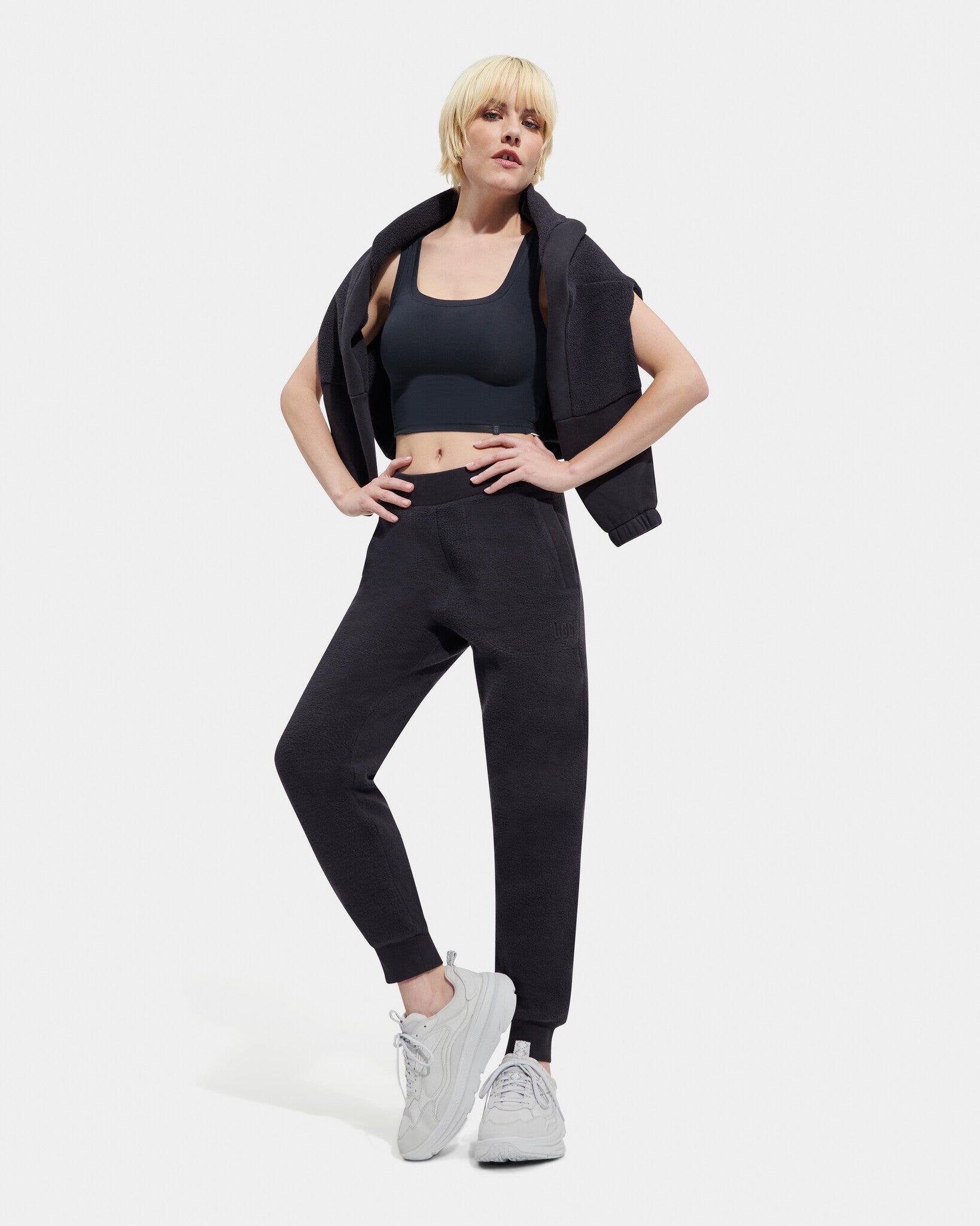 A female model standing, posing, and turning to face the camera. She is wearing a black UGG Ink Sofiana Mixed Jogger with an elastic waistband, and white sneakers. Available at Be Charmed