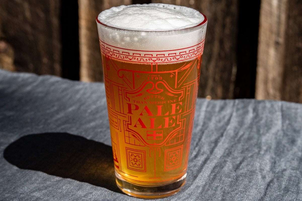 Pint glass of pale ale with decorative text on a wooden surface. Available at Be Charmed