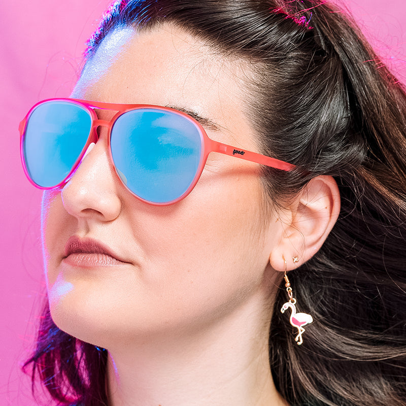 A person wearing pink and teal sunglasses with a reflective mirrored lens, standing against a pink background. Available at Be Charmed