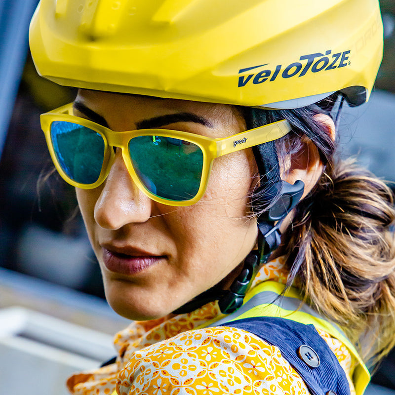 A person wearing yellow and blue Goodr sunglasses with blue reflective lenses and a black helmet with the logo 'velotezze' on it. Available at Be Charmed
