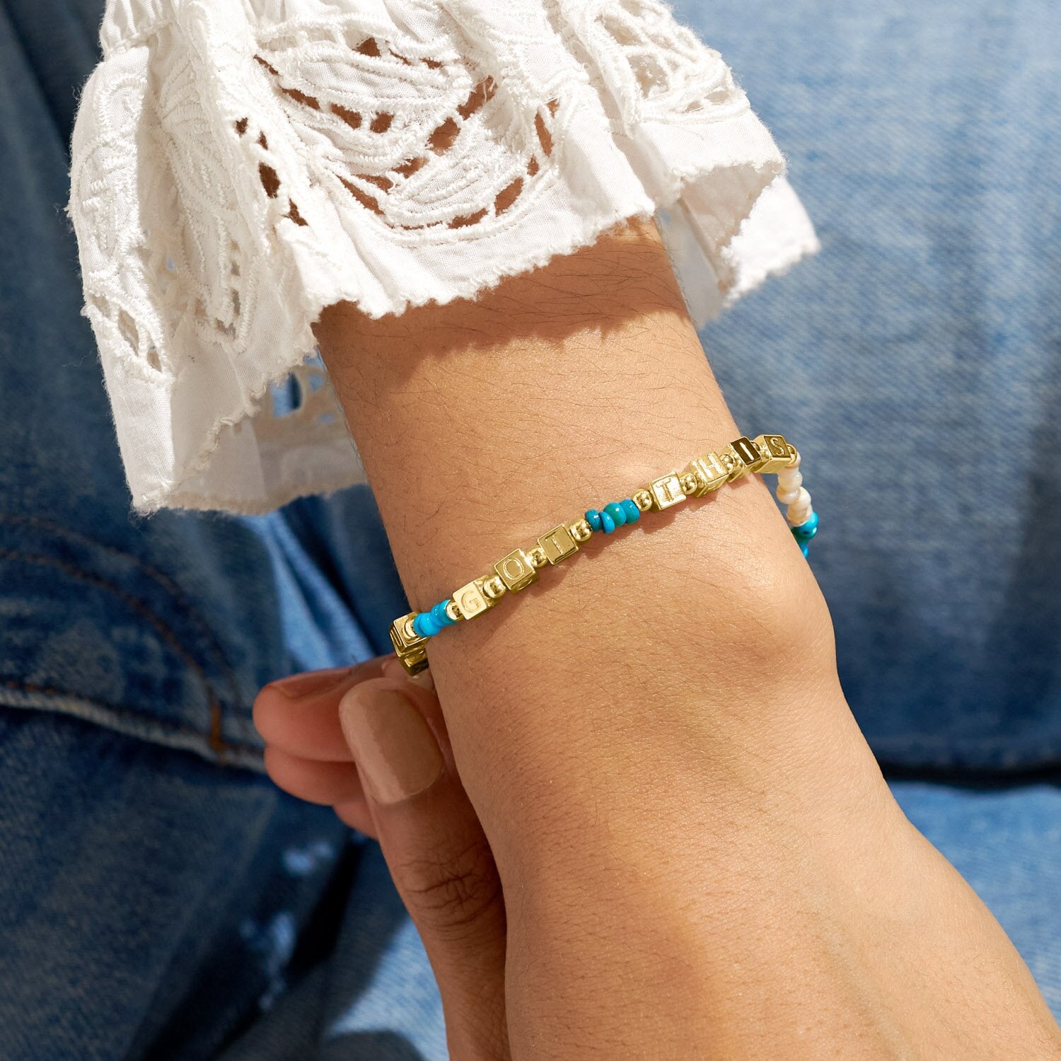 Close-up of a wrist wearing a gold and blue beaded bracelet against a blue background. Available at Be Charmed