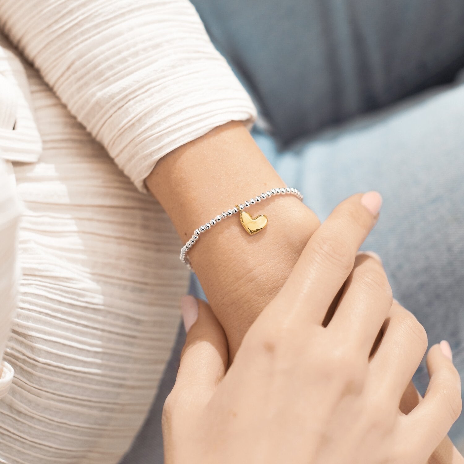 Close-up of a hand wearing a gold heart-shaped bracelet on a blurred background. Available at Be Charmed