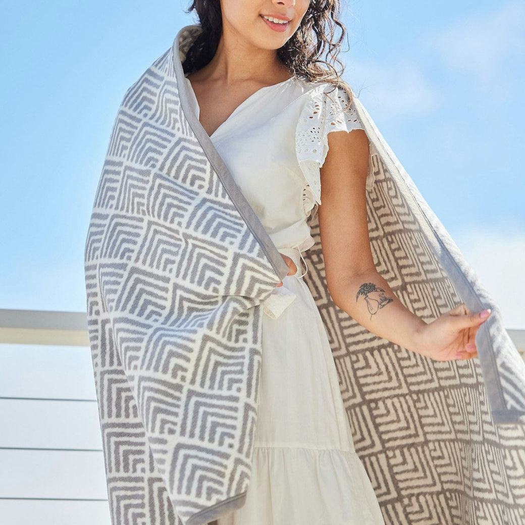 Woman wrapped in Oyster Cove Grey ChappyWrap Blanket featuring a classic diamond weave pattern, made from a cotton-blend fabric for exceptional softness and durability—ideal for cozy nights or elegant home décor. Available at Be Charmed.