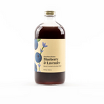 A bottle labeled 'Wood Stove Kitchen Blueberry & Lavender' with a clear liquid visible through the bottle, set against a white background. Available at Be Charmed