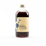 A bottle labeled 'Wood Stove Kitchen Blueberry & Lavender' with a clear liquid visible through the bottle, set against a white background. Available at Be Charmed