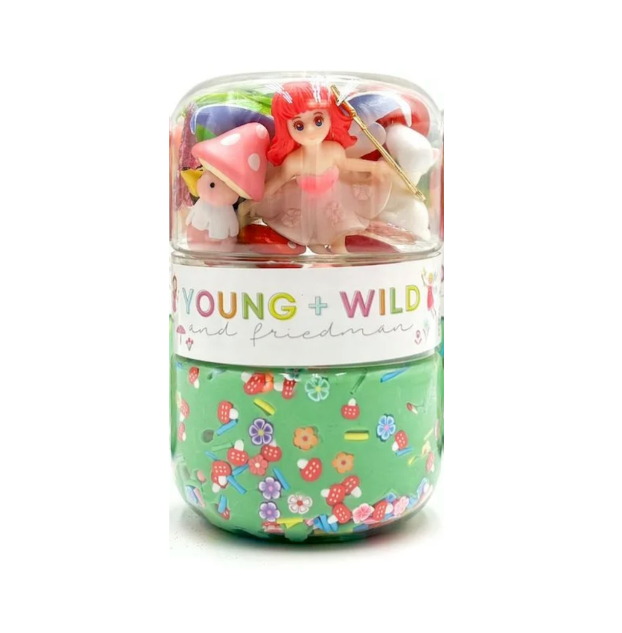 A clear cylindrical capsule-style container with a white label reading 'YOUNG + WILD and friedman' and a green lower section covered in small multicolor candy-like illustrations. Inside the capsule are small figurines, including a red-haired character and a white character with a pink hat, all on a plain white background. Available at Be Charmed