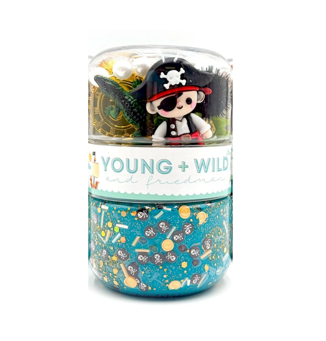 A clear cylindrical container with a white label reading 'YOUNG + WILD and friendship' holding a colorful pirate-themed craft or toy set. The top half is transparent and shows a small cartoon pirate figure, a black eye patch, a gold coin, and other small accessories. Available at Be Charmed
