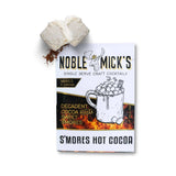 Noble Mick’s single-serve S’mores Hot Cocoa packet with a marshmallow-topped mug illustration, pictured beside marshmallows. Available at Be Charmed