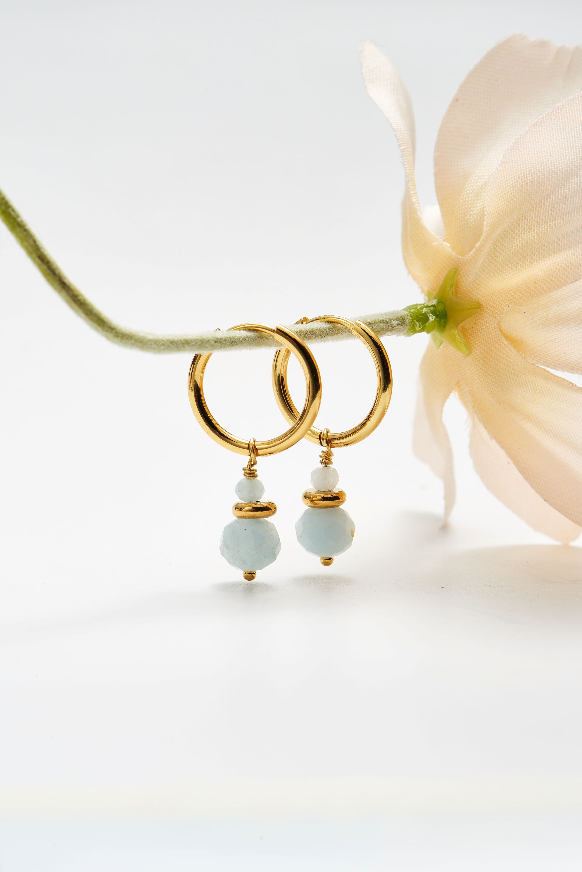 Gold hoop earrings with light blue bead drops, styled with a pale flower stem on a clean white background.