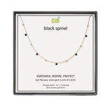 A CAI gold-plated necklace with small black gemstone beads displayed on a white card inside a black gift box. The card shows the brand name 'cai' and the text 'black spinel'. Available at Be Charmed