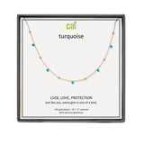 A CAI gold-plated necklace with small turquoise gemstones displayed on a branded white card inside a black presentation box. The card reads 'cai turquoise' and 'LUCK, LOVE, PROTECTION'. Available at Be Charmed