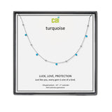 A CAI silver-toned necklace with small turquoise gemstones displayed on a white presentation card inside a black gift box. The card shows the brand name 'cai' and the word 'turquoise', along with the text 'LUCK, LOVE, PROTECTION'. Available at Be Charmed