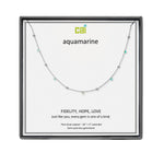 A CAI silver necklace with small aquamarine gemstones displayed on a white card inside a black square gift box. The card shows the brand name 'cai' and the word 'aquamarine' with additional product text below. Available at Be Charmed