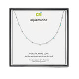 A CAI silver necklace with small aquamarine gemstones displayed on a white card inside a black square gift box. The card shows the brand name 'cai' and the word 'aquamarine' with additional product text below. Available at Be Charmed