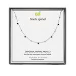 A CAI silver-toned necklace with small black gemstones displayed on a branded white card inside a dark gray square presentation box. The card reads 'cai black spinel' and includes product text below. Available at Be Charmed