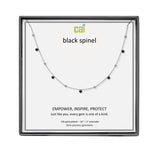 A CAI silver-toned necklace with small black gemstones displayed on a branded white card inside a dark gray square presentation box. The card reads 'cai black spinel' and includes product text below. Available at Be Charmed
