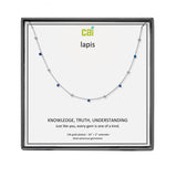 A CAI silver necklace with small blue and clear gemstones displayed on a white card inside a square black gift box. The card is branded with the logo 'cai' and the word 'lapis'. Available at Be Charmed