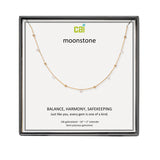 A CAI gold-plated necklace with small clear gemstones displayed on a white card inside a black square gift box. The card shows the brand name 'cai' and the product name 'moonstone'. Available at Be Charmed