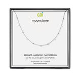 A CAI silver necklace with small spaced beads displayed on a white card inside a square black gift box. The card shows the brand name 'cai' and the product name 'moonstone', along with descriptive text. Available at Be Charmed