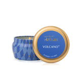 A blue and gold scented candle tin shown open with the lid leaning in front. The lid features a blue label with gold and white text reading 'capri BLUE Artisan VOLCANO'. Available at Be Charmed