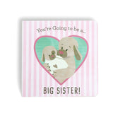 A book with a pink and white striped background featuring illustrations of two dogs and a baby, with the text 'You're Going To Be a Big Sister!' and 'Big Brother!'. Available at Be Charmed