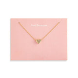 A delicate Demdaco gold necklace with a small heart-shaped pendant, presented on a light pink display card that reads 'Just Because.' against a clean white background. Available at Be Charmed