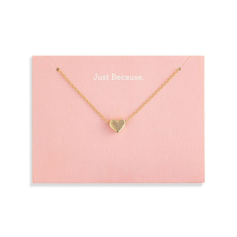 A delicate Demdaco gold necklace with a small heart-shaped pendant, presented on a light pink display card that reads 'Just Because.' against a clean white background. Available at Be Charmed
