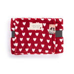A folded Demdaco red plush blanket with a white heart pattern, featuring a small cream label and a decorative tag on the front, set against a plain white background. Available at Be Charmed