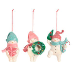 Three Demdaco Christmas snowman ornaments on a white background, each with a red hanging string. One snowman holds a pink gift box, one holds a green glittery wreath, and one holds a small green tree and a blue scarf. Available at Be Charmed