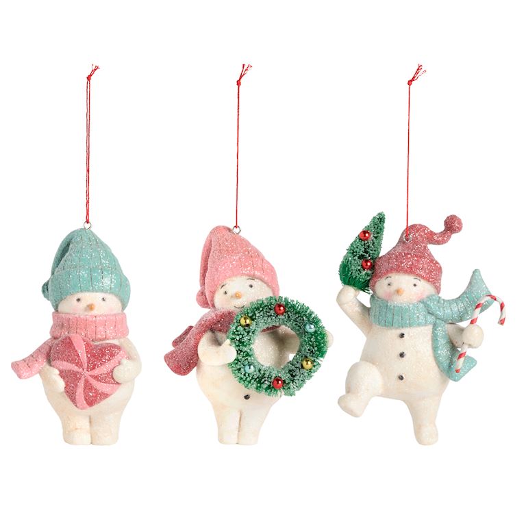 Three Demdaco Christmas snowman ornaments on a white background, each with a red hanging string. One snowman holds a pink gift box, one holds a green glittery wreath, and one holds a small green tree and a blue scarf. Available at Be Charmed
