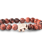 A beaded stretch fahlo bracelet made of round reddish-brown and black marbled beads with a small carved off-white elephant charm in the center, shown on a white background. Available at Be Charmed