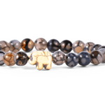 A beaded stretch bracelet featuring polished round beads in smoky gray, black, and brown marbled patterns, with a small elephant charm at the front center, set against a plain white background. Available at Be Charmed