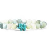 A beaded stretch fahlo bracelet featuring light green, white, and pale blue-green round beads with a small turquoise elephant charm in the center, set against a plain white background. Available at Be Charmed