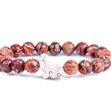 A beaded stretch Fahlo bracelet featuring round reddish-brown and black marbled beads with a small white ceramic lion charm in the center, displayed on a clean white background. Available at Be Charmed